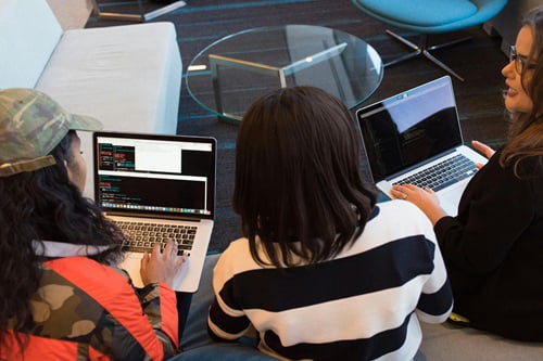 Three women are seated on the floor in a modern office, each working on a laptop displaying code or terminal windows.
