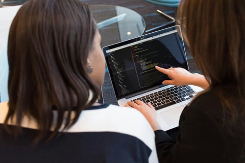 Two women are collaborating, looking at a laptop screen displaying lines of code. One woman, seen from behind with dark hair, is typing on the keyboard, while the other woman, also with dark hair, points to a specific section of the code on the screen.