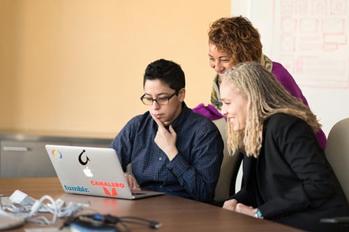 Three colleagues collaborate around a laptop on a conference table, with one person intently looking at the screen while two others smile and observe.