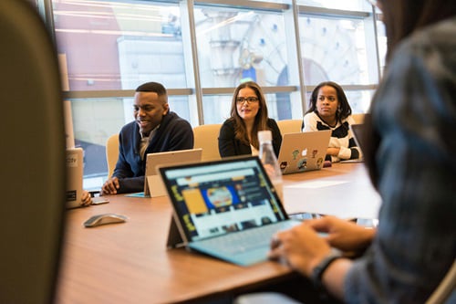 A diverse group of professionals attend a meeting in a bright conference room, with some looking at laptops and others engaged in discussion.