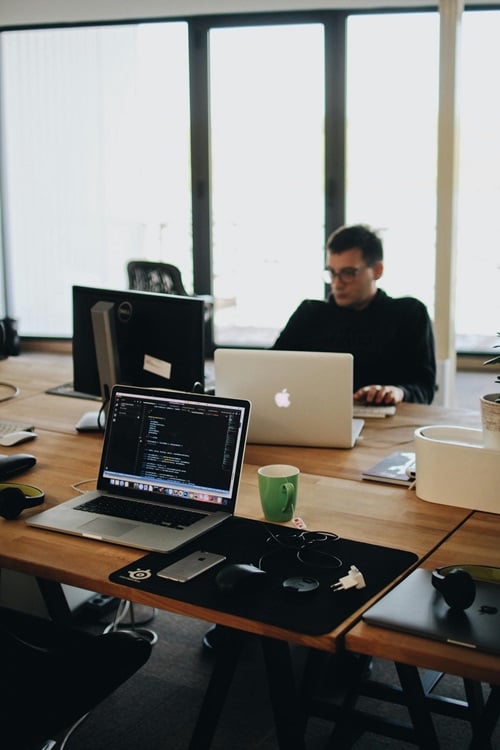 A man in glasses works on a laptop at a wooden desk, with another laptop displaying code in the foreground, alongside a green mug and a smartphone.