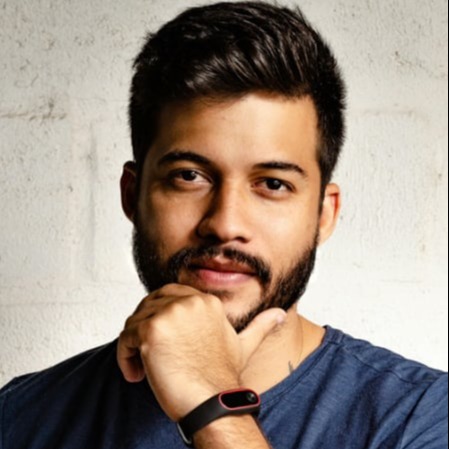 A man with dark hair and a beard, wearing a blue t-shirt, with his right hand on his chin, looks directly forward. He is wearing a black fitness tracker with a red accent on his left wrist. The background is a textured white wall.