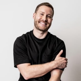 A smiling man with a beard, wearing a black t-shirt and light-colored pants, sits on a stool with his arms crossed. He is wearing a black smartwatch on his left wrist, against a white background.
