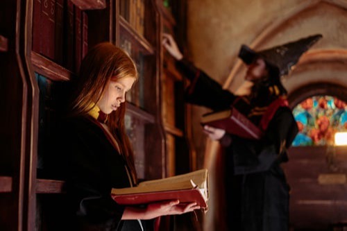 A young person with reddish-brown hair, dressed in a dark robe with yellow and red accents, reads a thick book in a dimly lit, old library. In the background, another person, wearing a wizard's hat, reaches for a book on a higher shelf, next to a stained-glass window.