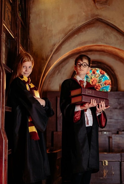 Two children dressed in wizarding robes stand in a magical library. A girl with long red hair, wearing a black robe and a yellow and red striped scarf, smiles while leaning against a bookshelf on the left. A boy with glasses, wearing a black robe and a red and yellow tie, holds a stack of old books and looks forward on the right. In the background, there's an arched ceiling and a stained-glass window.