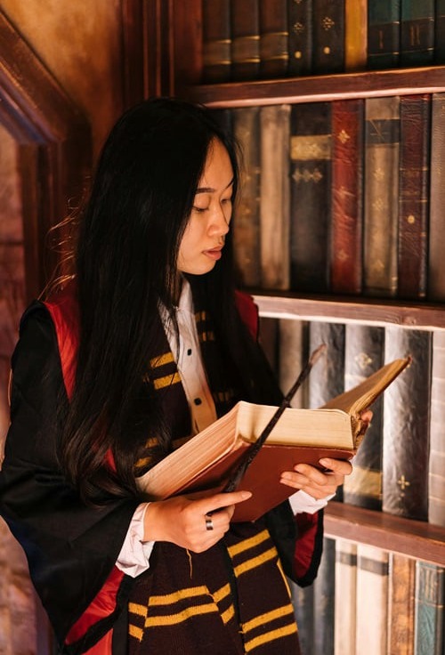 A young woman with long dark hair, dressed in a black and red wizarding robe and a yellow and dark striped scarf, intently reads a large, old book. A wand is tucked into the pages of the book, and she is surrounded by shelves filled with many old books in a library setting.